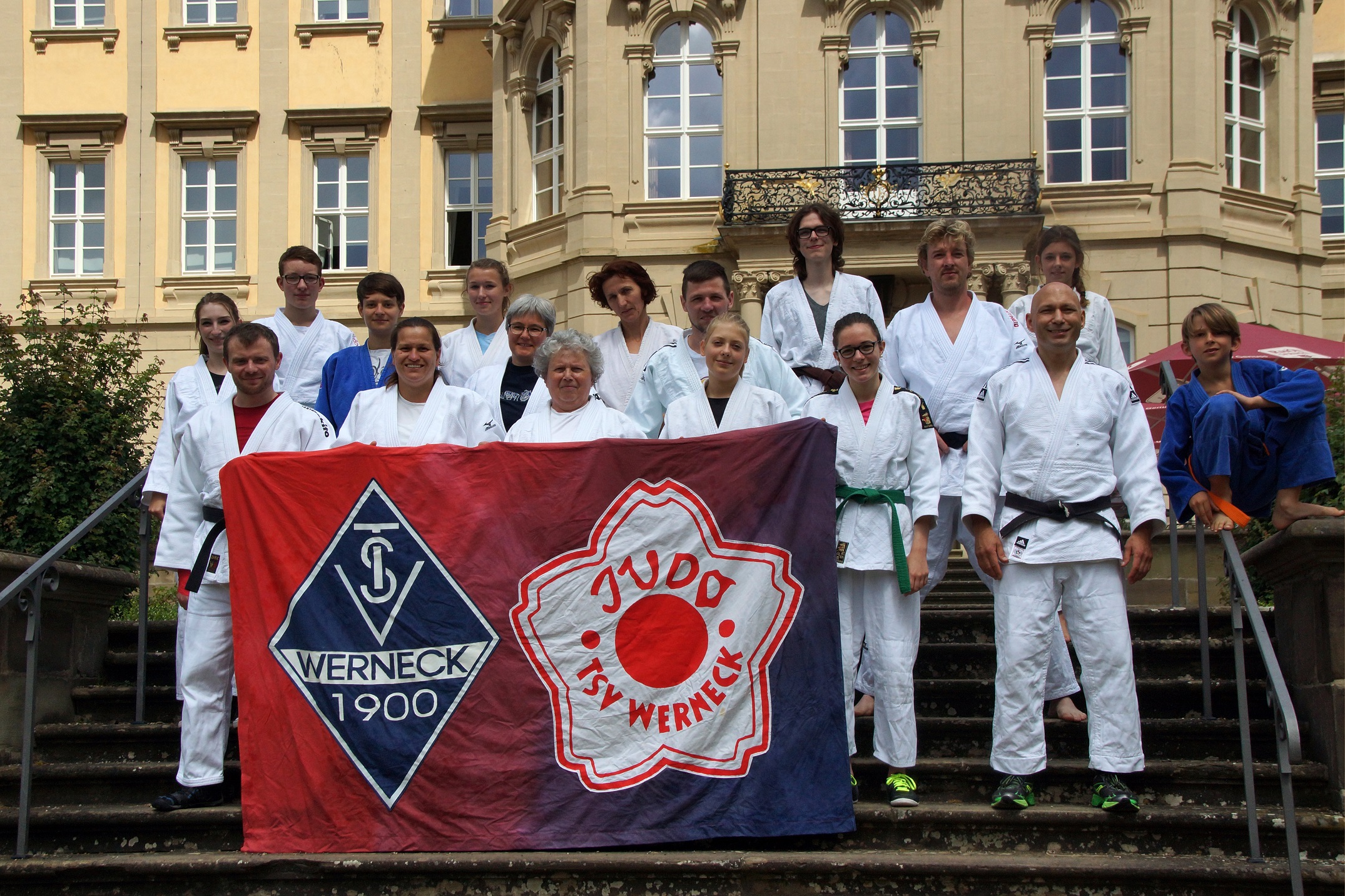 Bild: Gruppen Bild, alle Judokas haben ihren Judoanzug an. Es wird ein Poster nach vorn gehalten auf dem das Logo des Sportvereins und der Judo Abteilung ist.