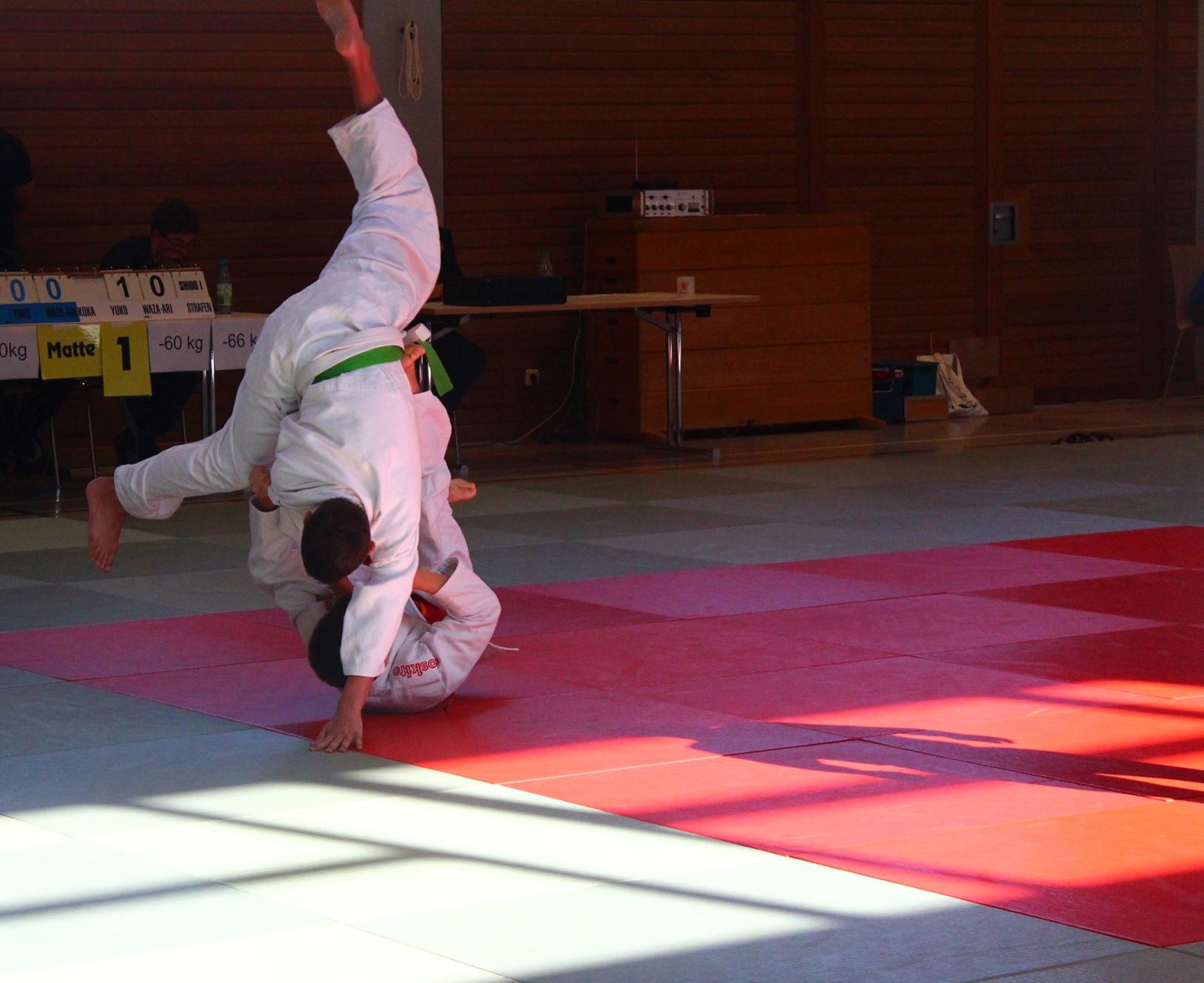 Bild von einem Wettkampf, ein Judoka mit Grünen Gürtel fällt über ein am Boden liegendes Judoka und Stützt dabei seine Hand auf der Matte ab. 